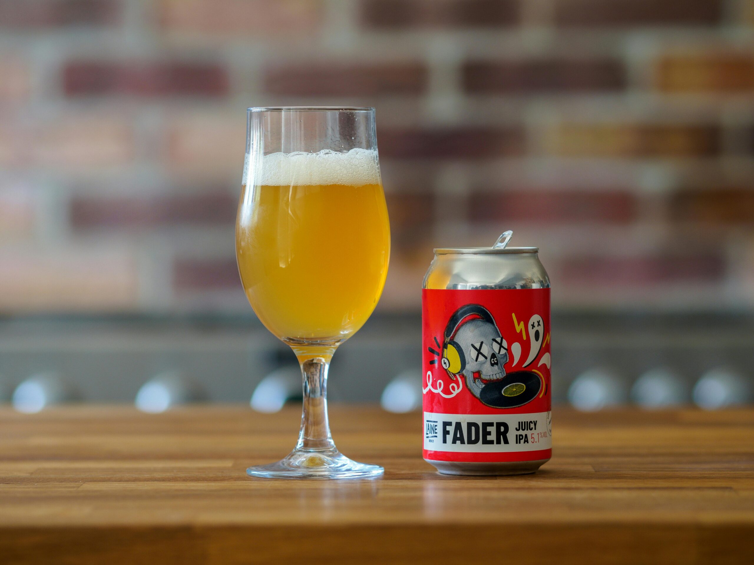 A vibrant juicy IPA in a glass and can displayed on a wooden bar with a blurred brick background.